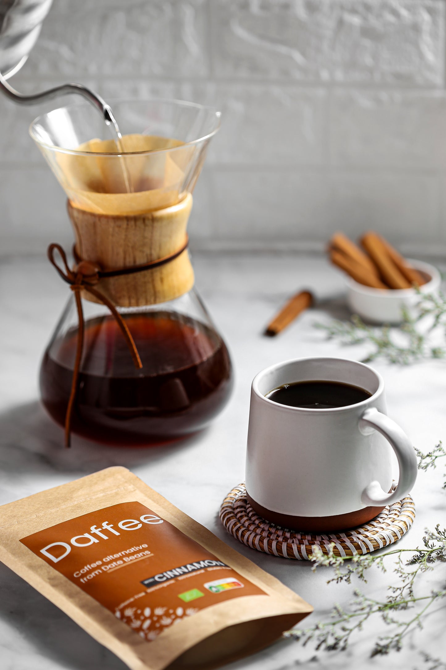 Een karaf met Daffee koffie naast een mok op een onderzetter en een Daffee Cinnamon pakket, met een bloeiende tak op de achtergrond